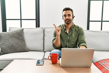 Young hispanic man with beard wearing call center agent headset working from home pointing with hand finger to the side showing advertisement, serious and calm face