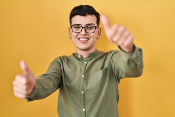 Non binary person standing over yellow background approving doing positive gesture with hand, thumbs up smiling and happy for success. winner gesture.