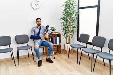 Young arab man sitting on chair with arm sling holding crutch at clinic waiting room