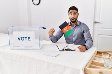 Young handsome man with beard at political campaign election holding afghanistan flag scared and amazed with open mouth for surprise, disbelief face