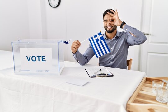 Young handsome man with beard at political campaign election holding greece flag smiling happy doing ok sign with hand on eye looking through fingers