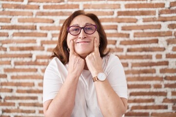 Senior woman with glasses standing over bricks wall smiling with open mouth, fingers pointing and forcing cheerful smile