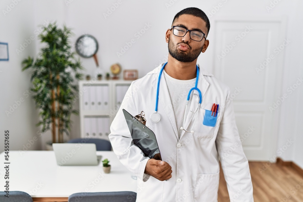 Wall mural Young indian man wearing doctor uniform and stethoscope looking at the camera blowing a kiss on air being lovely and sexy. love expression.