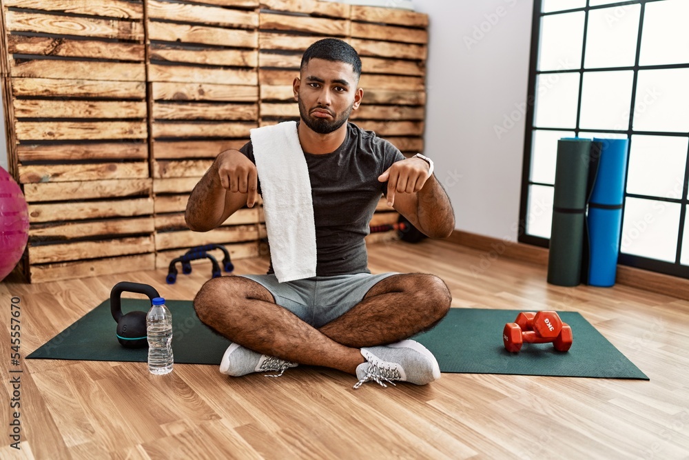 Sticker young indian man sitting on training mat at the gym pointing down looking sad and upset, indicating 