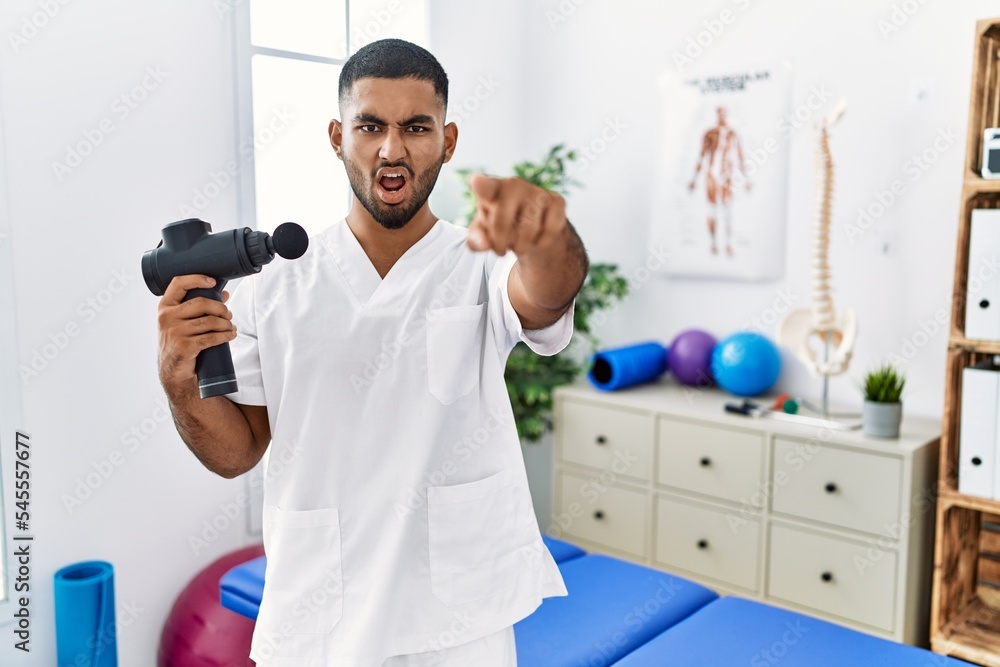 Sticker young indian physiotherapist holding therapy massage gun at wellness center pointing displeased and 