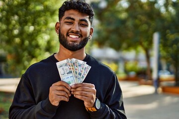 Young arab man smiling confident holding sol peruvian banknotes at park - obrazy, fototapety, plakaty