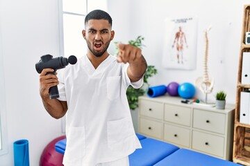 Young indian physiotherapist holding therapy massage gun at wellness center pointing displeased and...