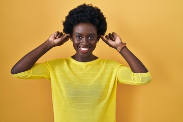 African young woman standing over yellow studio smiling pulling ears with fingers, funny gesture. audition problem