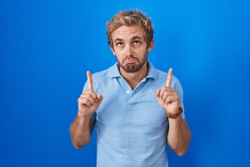 Caucasian man standing over blue background pointing up looking sad and upset, indicating direction...