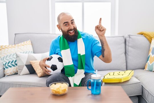 Young hispanic man with beard and tattoos football hooligan holding ball supporting team smiling happy pointing with hand and finger to the side