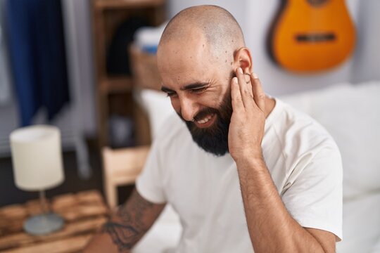Young bald man suffering for ear pain sitting on bed at bedroom