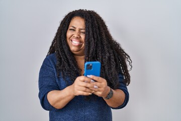 Plus size hispanic woman using smartphone typing message winking looking at the camera with sexy...
