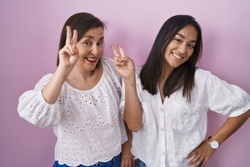 Hispanic mother and daughter together smiling looking to the camera showing fingers doing victory sign. number two.