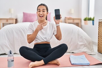 Fototapeta na wymiar Young south asian woman doing yoga mat holding smartphone showing screen smiling happy pointing with hand and finger