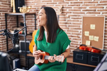 Young hispanic woman musician singing song playing ukulele at music studio