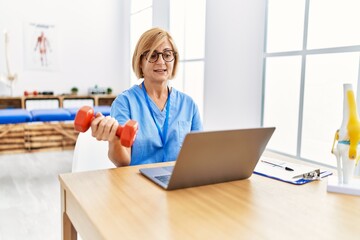 Middle age blonde woman wearing physio therapy uniform doing tele rehab using dumbbells at clinic