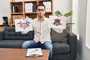 Young hispanic man with beard holding rorschach test making fish face with mouth and squinting eyes, crazy and comical.