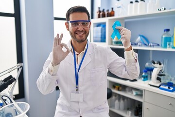 Young hispanic man with beard working at scientist laboratory holding blue ribbon doing ok sign...