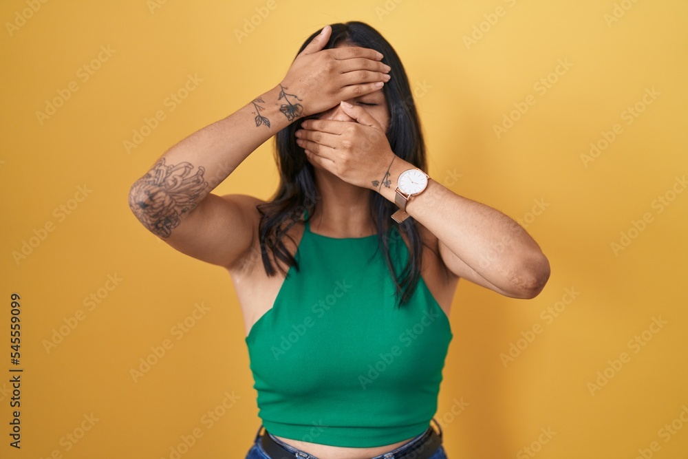 Poster brunette woman standing over yellow background covering eyes and mouth with hands, surprised and sho