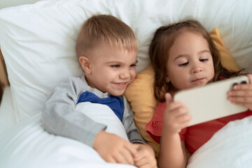 Fototapeta na wymiar Adorable girl and boy watching video on smartphone lying on bed at bedroom