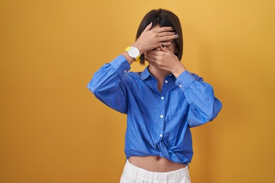 Young girl standing over yellow background covering eyes and mouth with hands, surprised and shocked. hiding emotion