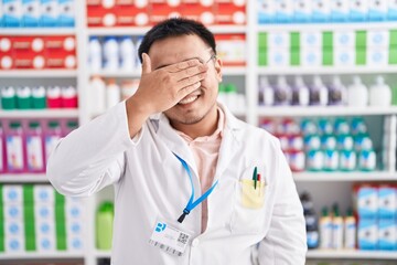 Chinese young man working at pharmacy drugstore smiling and laughing with hand on face covering eyes for surprise. blind concept.