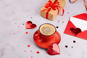 A red cup of coffee with milk, a gift in kraft paper, red envelope with an empty sheet of paper, heart decorations on a gray background. A Valentine's Day card. 