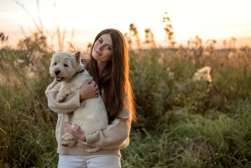 portrait of a beautiful young girl with a dog in her arms, a girl hugs her beloved petam on a walk at the sunset, West Highland White Terrier, copy space