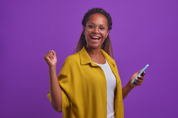 Young joyful beautiful African American woman zoomer with phone in hand makes victorious wave of hand and shouts rejoicing at passage of level in mobile game stands on lilac studio background