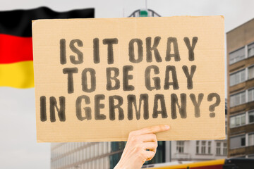 The question " Is it okay to be gay in Germany? " is on a banner in men's hands with blurred background. Passionate. Contact. Date. Dating. Lover. Partner. Boyfriend. Pleasant. Lovely. Okay. Approval