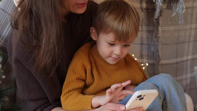 A cute little boy and his mom are reading a fairy tale on a mobile phone, comfortably ensconced in a tent in a dark night bedroom. The concept of love, childhood, friendship, imagination, creativity