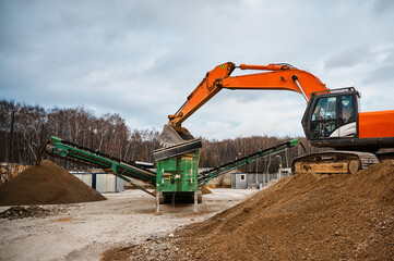 A construction excavator loads crushed stone into the receiving hopper of a mobile crushing and...