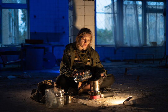 Post-apocalypse. a young woman sits in an abandoned building on the floor pours water into a cup for cooking. concept of a man hiking in a post-apocalyptic world looking for food.