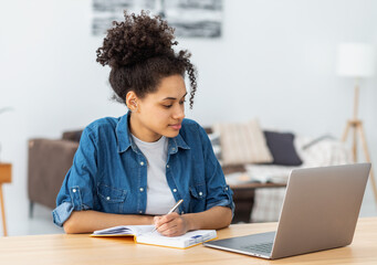 Concentrated young African American woman student using laptop computer for online education, taking notes in a notebook, studying, distance learning at home