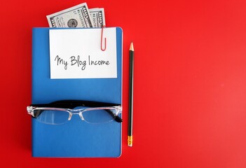 Reading Glasses, pencil, cash dollars money in blue notebook with text written note MY BLOG INCOME, on copy space red background, concept of blogger make more income earnings cash money from blogging 