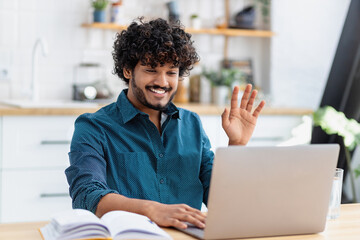 Young happy Asian man freelancer or entrepreneur have video conference, talking, shows a greeting gesture, working remotely online at home office