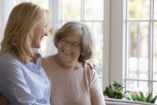 Caring middle-aged woman hugging her senior grey-haired mother in glasses standing together at home laughing enjoy positive friendly talk and time together. Multi-generational family, ties and love