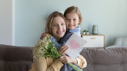 Portrait of smiling young mother and little daughter sitting on couch look at camera celebrating birthday together, loving girl congratulate happy mom with anniversary embracing hold flowers and card
