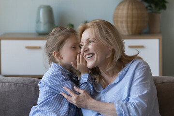 Cute little girl whisper in ear tell secret to cheerful grandma. Happy middle-aged woman her granddaughter enjoy their friendly relations, trust and understanding, talking at home, spend time together