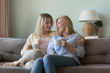 Understanding, harmonic relationships between older mother and grown up millennial daughter. Two different generation female sit on couch with teacups smile enjoy pleasant talk and warm communication