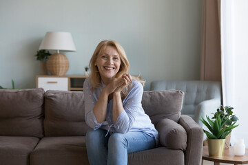 Pretty middle-aged woman posing for picture seated on sofa in modern living room. Portrait of happy, good-looking optimistic female spend carefree leisure at home, enjoy untroubled life on retirement - obrazy, fototapety, plakaty