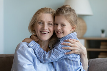 Portrait of grandmother hugs her cute little granddaughter sit on sofa smile staring at camera...
