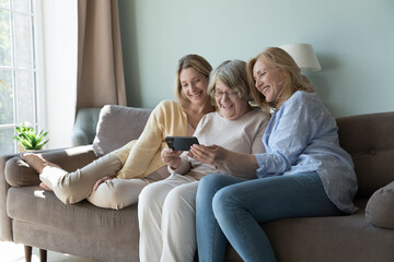 Obraz premium Caring younger generation teach older gen to use modern wireless technology, showing new mobile app for comfort life, spend carefree leisure sit together on sofa at home staring at smartphone screen