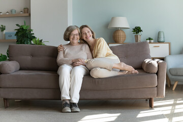 Obraz premium Young adult granddaughter hugs joyful retired older grandmother relax on sofa smile look at camera. Portrait of happy different generations family, showing care, appreciate time together, feeling love
