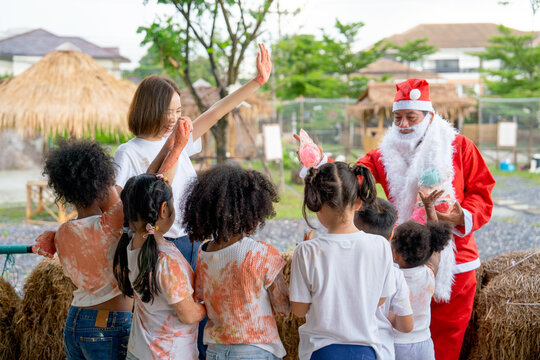 Santa Claus man give many gifts or present to group of multi-ethnic children after they enjoy to play with color paint with teacher during Christmas celebration.