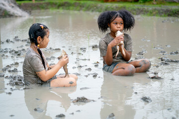 African American girl sit and kiss frog during fun play in mud playground with Asian friend and...