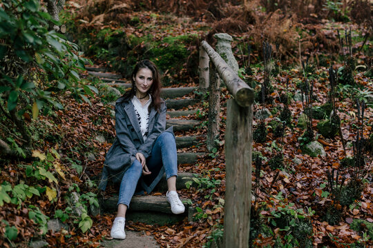 A beautiful brunette woman in her 30s sits on old wooden steps in an autumn park. Yellow-orange colorful autumn. Blurred background.
