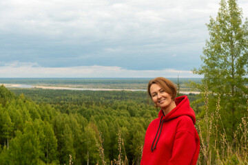 a woman with red hair and a red hoodie on a wooded hill against the background of a forest and a winding river in the distance.