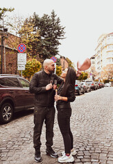 Obraz premium Romantic couple in casual outfits toasting with champagne outdoors and posing on streets of european city. Beautiful woman clinking with her husband.Happy couple with a glass of wine celebrating 