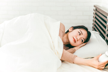 ベッドで眠くて寝ぼけながら目覚まし時計を止める低血圧の日本人女性
